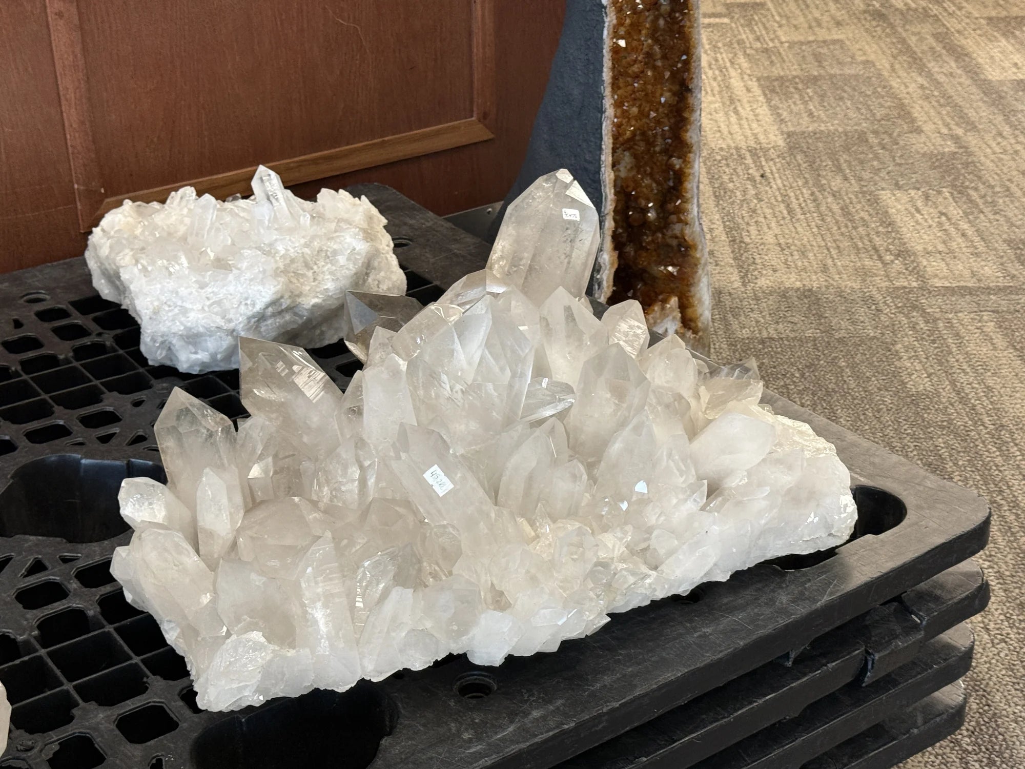A large clear quartz crystal cluster on a black plastic crate, with a smaller white mineral specimen and a vertical brown crystal specimen in the background, set on a carpeted floor.