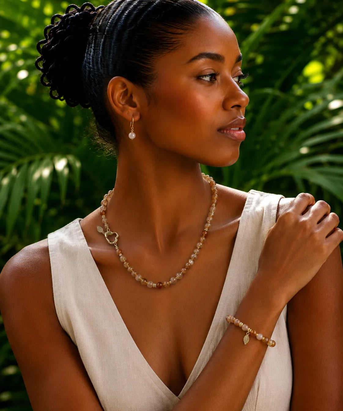 A woman wearing a delicate citrine beaded necklace and matching bracelet with small leaf charms, styled with a white sleeveless top against a lush green tropical background.
