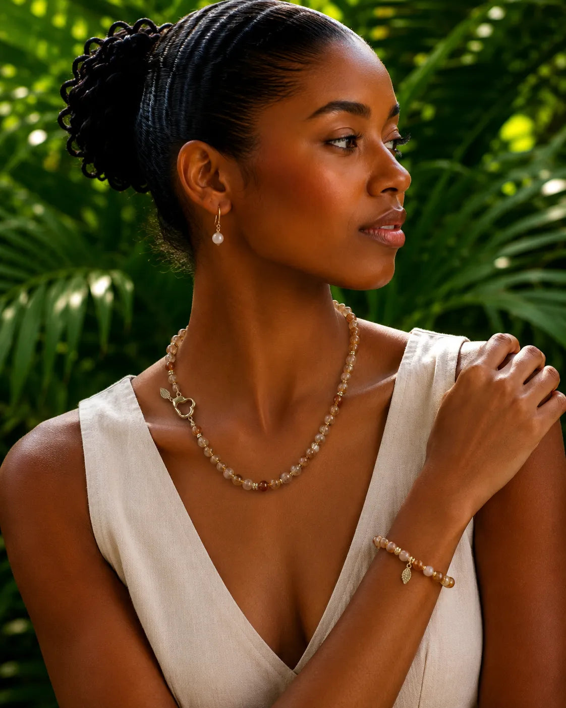 A woman wearing a delicate citrine beaded necklace and matching bracelet with small leaf charms, styled with a white sleeveless top against a lush green tropical background.