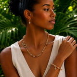 A woman wearing a delicate citrine beaded necklace and matching bracelet with small leaf charms, styled with a white sleeveless top against a lush green tropical background.