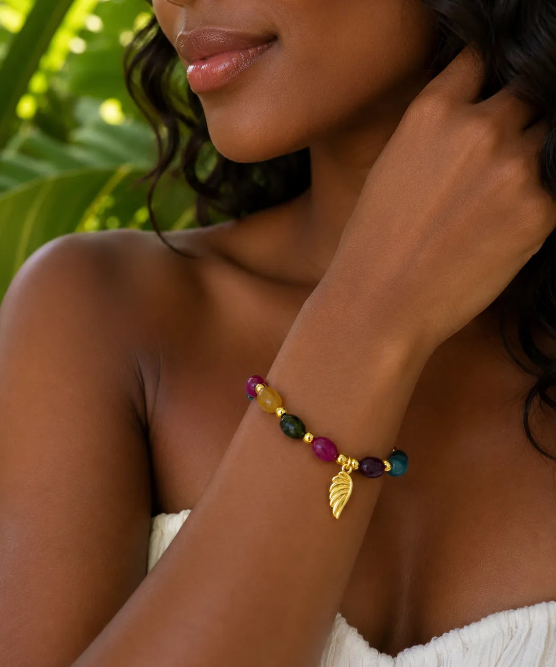 A close-up of a woman wearing a colorful beaded jade bracelet with a gold leaf charm, set against a background of green tropical leaves.