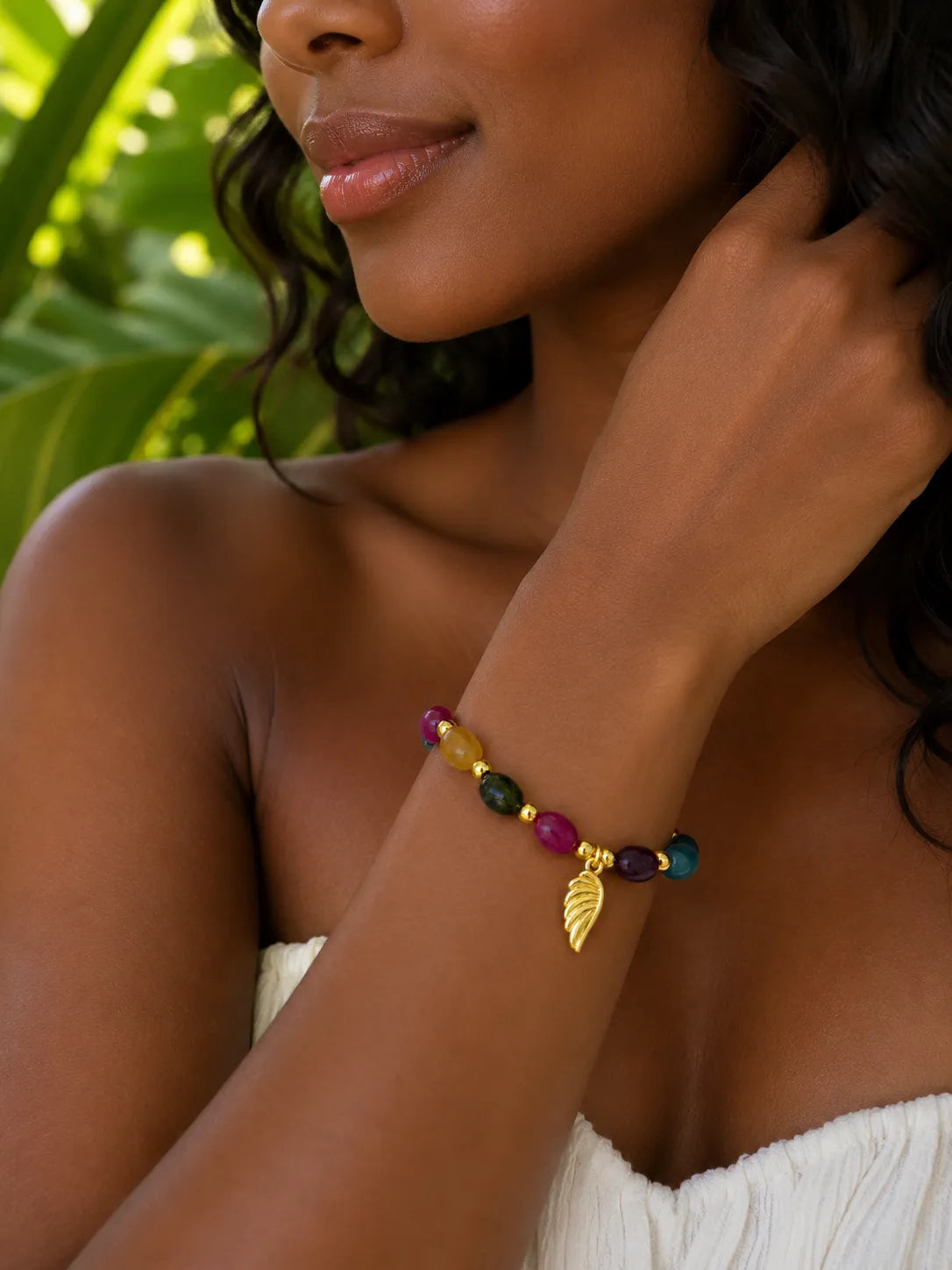 A close-up of a woman wearing a colorful beaded jade bracelet with a gold leaf charm, set against a background of green tropical leaves.