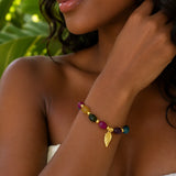 A close-up of a woman wearing a colorful beaded jade bracelet with a gold leaf charm, set against a background of green tropical leaves.