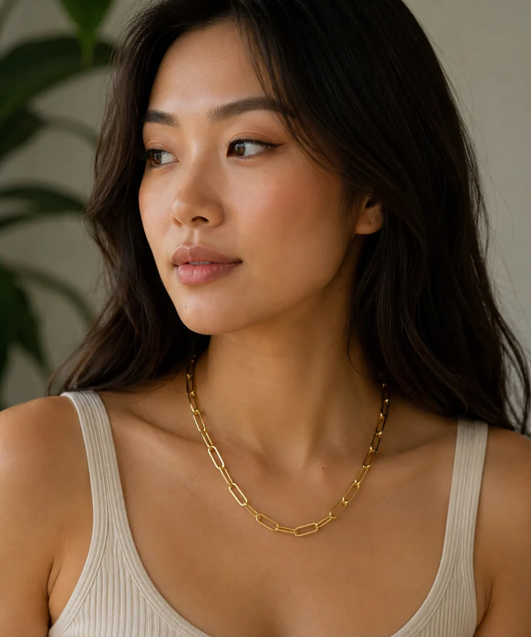 A woman with long dark hair wearing a gold paper clip chain necklace by MariaRyan Jewelry and a ribbed beige tank top, shown in a close-up portrait against a soft neutral background with a blurred green plant.