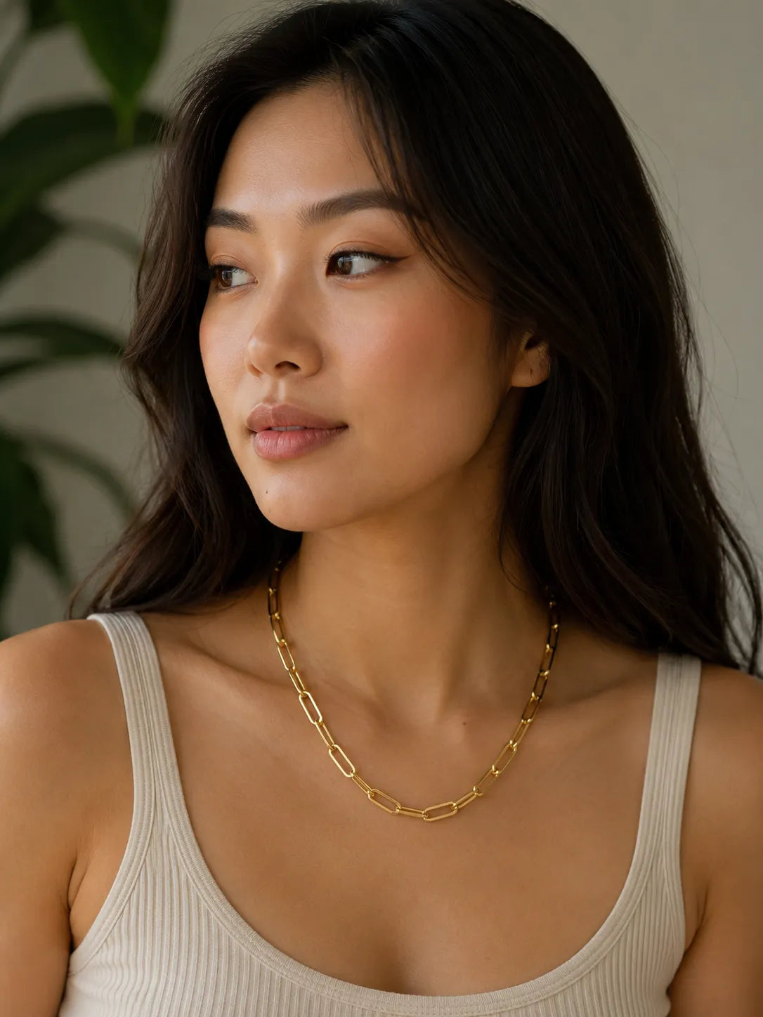 A woman with long dark hair wearing a gold paper clip chain necklace by MariaRyan Jewelry and a ribbed beige tank top, shown in a close-up portrait against a soft neutral background with a blurred green plant.