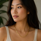 A woman with long dark hair wearing a gold paper clip chain necklace by MariaRyan Jewelry and a ribbed beige tank top, shown in a close-up portrait against a soft neutral background with a blurred green plant.