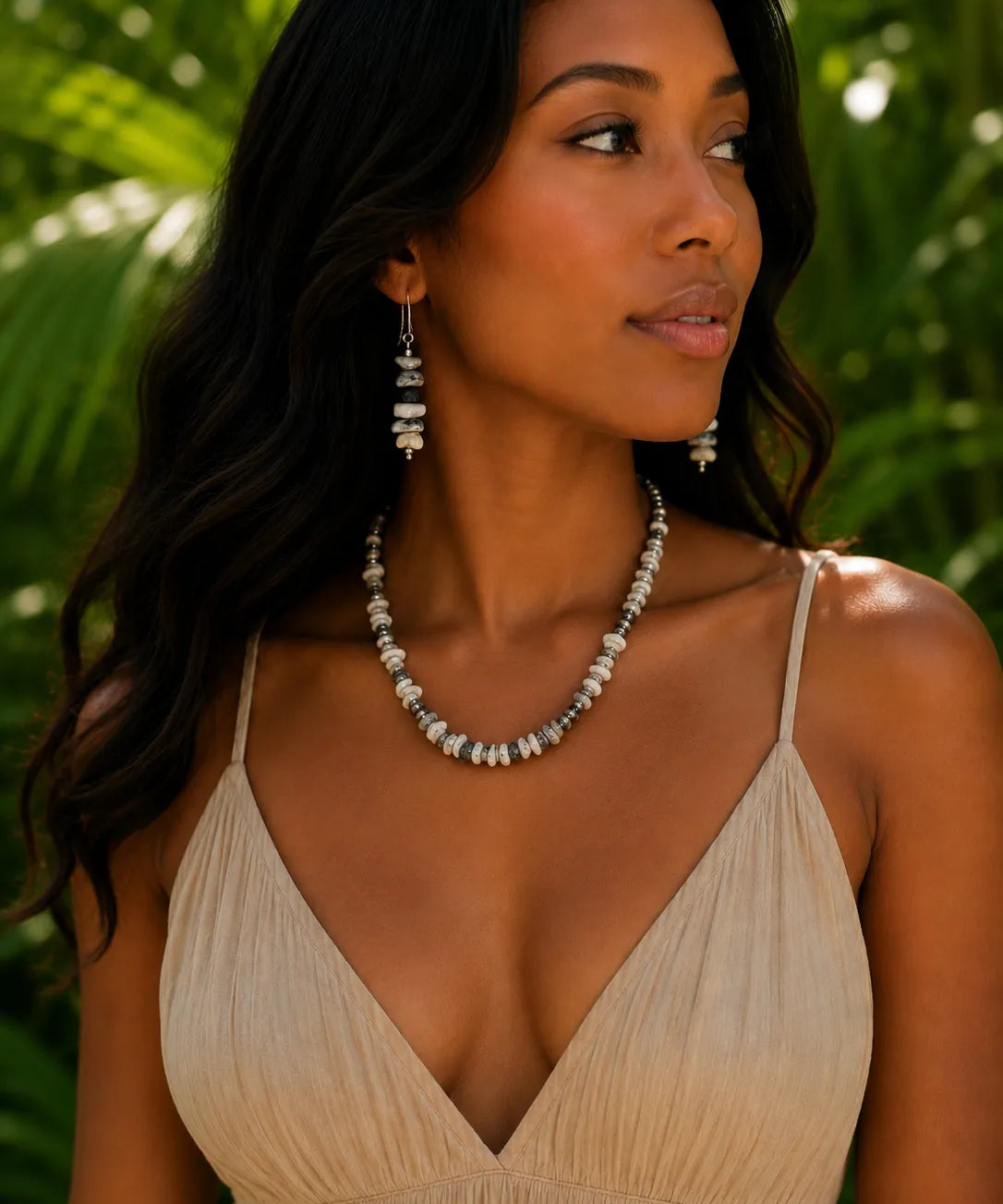 A woman wearing a silver-toned buffalo beaded necklace and matching drop earrings, styled with a beige spaghetti-strap dress, set against a blurred tropical green foliage background.