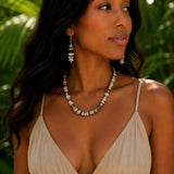 A woman wearing a silver-toned buffalo beaded necklace and matching drop earrings, styled with a beige spaghetti-strap dress, set against a blurred tropical green foliage background.